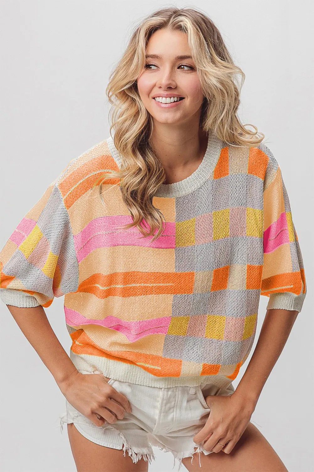 A smiling woman with beautiful wavy hair wearing a colourful knit top with white denim shorts.